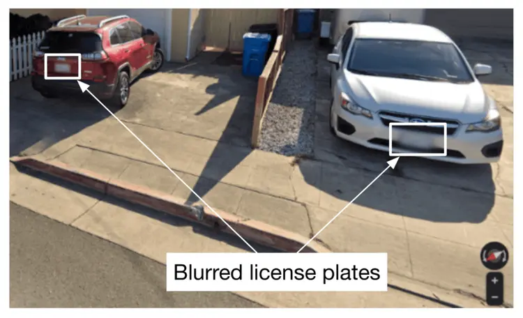 Image represents a street-level view of a residential driveway with two cars parked.  A red SUV is parked on the left, and a white sedan is parked on the right.  Both license plates are rectangular areas that have been intentionally blurred, indicated by white rectangular boxes overlaid on the image.  White arrows point from these blurred license plate boxes to the text 'Blurred license plates' located at the bottom center of the image.  The driveway is made of concrete, and a section of curb and a metal beam are visible in the foreground.  A portion of a white picket fence is visible in the upper left corner, and a blue recycling bin is partially visible behind the white car.  In the bottom right corner, there's a small compass-like icon and plus/minus buttons, suggesting the image is from a map application or similar platform allowing for zoom functionality.