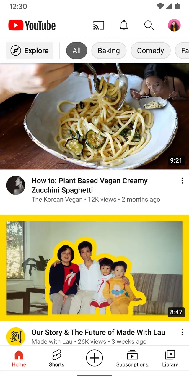 Image represents a screenshot of the YouTube mobile application.  At the top, a status bar displays the time '12:30' and standard mobile icons for Wi-Fi, notifications, search, and a user profile picture. Below this, the YouTube logo and title are prominent.  A horizontal navigation bar contains buttons for 'Explore,' 'All,' 'Baking,' 'Comedy,' and a partially visible fifth option. The main content area displays two video thumbnails. The top thumbnail shows a close-up of a bowl of zucchini spaghetti being eaten, with a video duration of '9:21' displayed in the corner.  Below the thumbnail, the video title 'How to: Plant Based Vegan Creamy Zucchini Spaghetti' is shown, along with channel information ('The Korean Vegan'), view count ('12K views'), and upload date ('2 months ago'). A three-dot menu icon is also present. The second thumbnail shows a family photo with a video duration of '8:47' and the title 'Our Story & The Future of Made With Lau,' along with channel information ('Made with Lau'), view count ('26K views'), and upload date ('3 weeks ago').  A three-dot menu icon is also present. At the bottom of the screen, a navigation bar with icons and labels for 'Home,' 'Shorts,' a plus icon (likely for creating content), 'Subscriptions,' and 'Library' is visible.  The overall layout is typical of a mobile app, with clear visual separation between navigation, content, and video information.